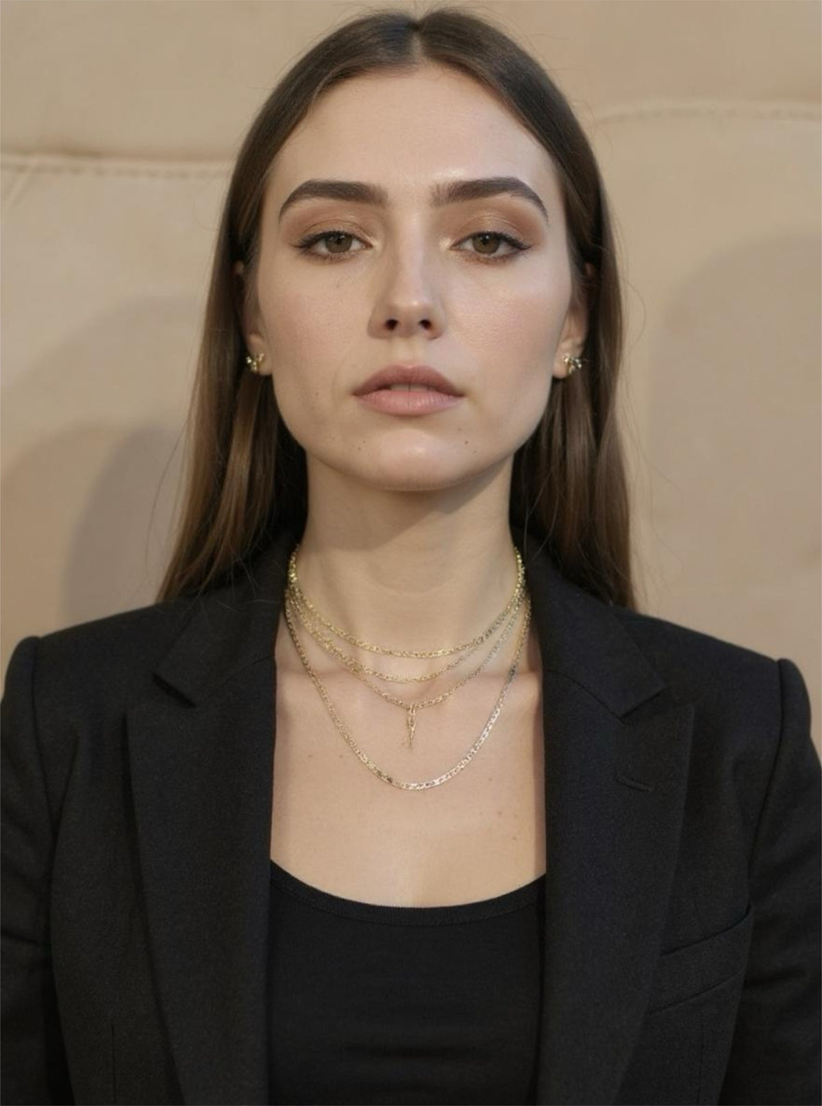 Woman wearing a black blazer and layered gold necklaces against a neutral background